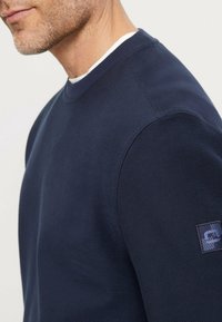 Marineblauwe sweatshirt met een ronde halslijn, geribde manchetten en een gestructureerde afwerking. Voorzien van een logopatch op de linkermouw.