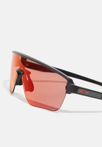 Sportsonnenbrille mit schwarzem Rahmen, großen rot getönten Gläsern und einem belüfteten Nasenstück. Mit eingraviertem Logo an der Seite.