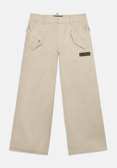 Pantalon beige à jambes larges avec poches à rabat à l'avant, fermetures à boutons, éclaboussures de peinture multicolores, et un patch de marque rectangulaire sur la cuisse droite.