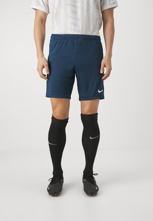 Mand iført blå atletikshorts, sorte knæstrømper og sorte fodboldstøvler, stående mod en hvid baggrund.