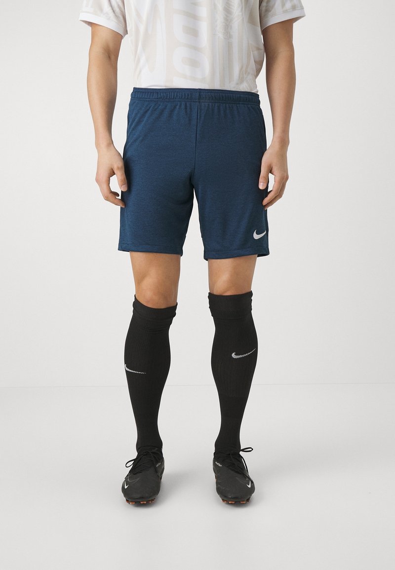 Homme portant un short de sport bleu, des chaussettes noires montantes et des chaussures de football noires, debout sur un fond blanc.
