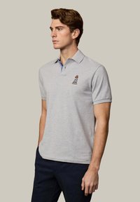 Uomo che indossa una polo a maniche corte grigio chiaro con un piccolo orso ricamato con maglia a righe e pantaloni scuri, che guarda a sinistra contro uno sfondo semplice.