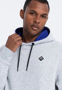 Sudadera gris con cordón, interior azul contrastante y logo de diamante negro en el pecho. Textura suave con puños acanalados.