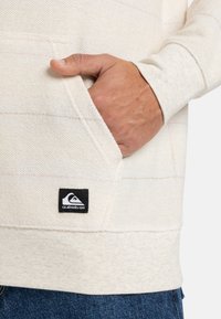 Lys beige hoodie med tekstureret stof, lange ærmer, fremadrettet lomme og et sort Quiksilver-logo mærke på kanten. Subtile farvestriber er til stede.
