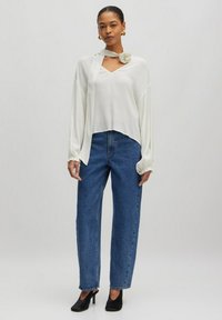Weißes, fließendes Langarmtop mit V-Ausschnitt und seitlichem Schal-Detail, kombiniert mit hoch taillierten, geraden blauen Jeans und schwarzen, hohen Pantoletten.