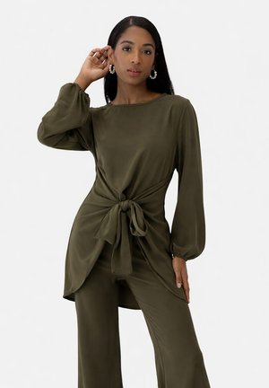 Femme aux longs cheveux foncés portant un haut à manches longues olive noué à la taille et un pantalon large assorti, accessoirisée avec des boucles d'oreilles créoles.