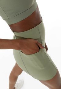 Persona vestida con pantalones cortos deportivos verde oliva y una camiseta, con una mano en el bolsillo, de pie con una pierna ligeramente doblada contra un fondo blanco.