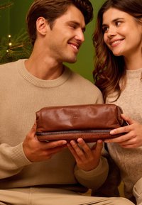 Bruine leren toilettas met ritssluiting, zachte textuur en subtiele branding, vastgehouden door twee personen tegen een groene achtergrond.