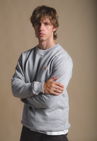 Sudadera gris de cuello redondo hecha de tela suave, con un pequeño logo de un elefante marrón en el lado izquierdo del pecho. El modelo está de pie con los brazos cruzados.