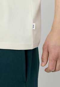 T-shirt beige à manches courtes en coton, avec une étiquette sur le côté portant "BZB". Assorti à un pantalon vert foncé, présentant un tissu texturé.