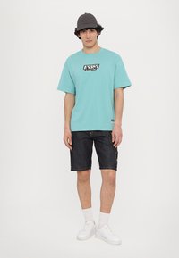 Τυρκουάζ κοντομάνικο μπλουζάκι με μαύρο λογότυπο, συνδυασμένο με σκούρες τζιν βερμούδες, λευκά αθλητικά παπούτσια και γκρι καπέλο, μπροστά σε λευκό φόντο.