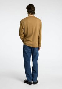 Ilga rankovė megztinis šiltos smėlio spalvos, laisvo kirpimo, derinamas su mėlynomis džinsinėmis kelnių. Audinys atrodo minkštas, su minimaliais siūlės, kad būtų patogu.