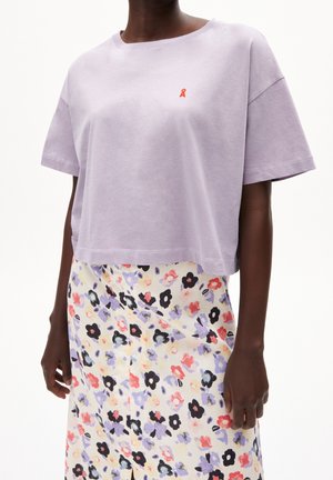 Kvinde iført en lyse lilla cropped T-shirt med lille rødt logo og en højtaljet cremefarvet nederdel med flerfarvet blomsterprint.