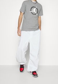 Hombre vestido con camiseta gris de Air Jordan, pantalones anchos blancos y zapatillas rojas, negras y blancas, de pie con una mano en el bolsillo.