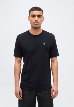 Muž stojaci čelom dopredu oblečený v čiernom tričku s malým vyšívaným logom na ľavej strane hrude a v čiernych nohaviciach na jednoduchom pozadí.