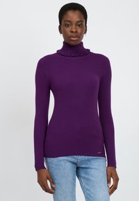 Lila gerippter Rollkragenpullover mit einem gerüschten Kragen, langen Ärmeln und tailliertem Schnitt, kombiniert mit hellblauen Jeans.