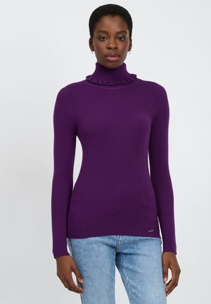 Lila gerippter Rollkragenpullover mit einem gerüschten Kragen, langen Ärmeln und tailliertem Schnitt, kombiniert mit hellblauen Jeans.