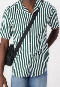 Camicia a maniche corte con bottoni, strisce verticali verdi e bianche, design con colletto, tessuto leggero, abbinata a una piccola borsa a tracolla nera.