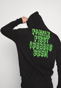 Person i en svart hoodie med neongrön text "FAMILY FIRST ICEBERG CREW" sedd bakifrån, lyfter en knuten näve nära huvudet.