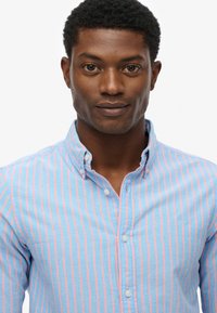 Jasnoniebieska koszula z długim rękawem w różowe paski, z kołnierzykiem i wyszywanym logo. Miękka faktura materiału, z klasycznym krojem.