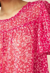 Blouse florale rose en tissu transparent, avec un col rond, des manches courtes et des bordures en dentelle délicate le long des bords et des coutures.
