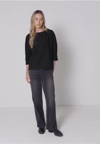 Zwarte driekwart mouwtop gecombineerd met grijze uitlopende jeans; model staat op een witte vloer, met steil haar en zwarte schoenen.