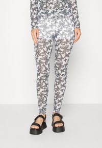 Leggings com padrão de borboletas em preto e branco feitos de tecido transparente, com um formato justo e faixa elástica na cintura. Combinados com sandálias pretas robustas.