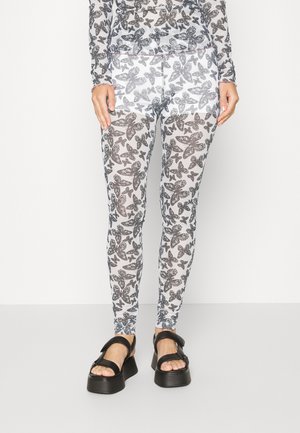 Leggings com padrão de borboletas em preto e branco feitos de tecido transparente, com um formato justo e faixa elástica na cintura. Combinados com sandálias pretas robustas.