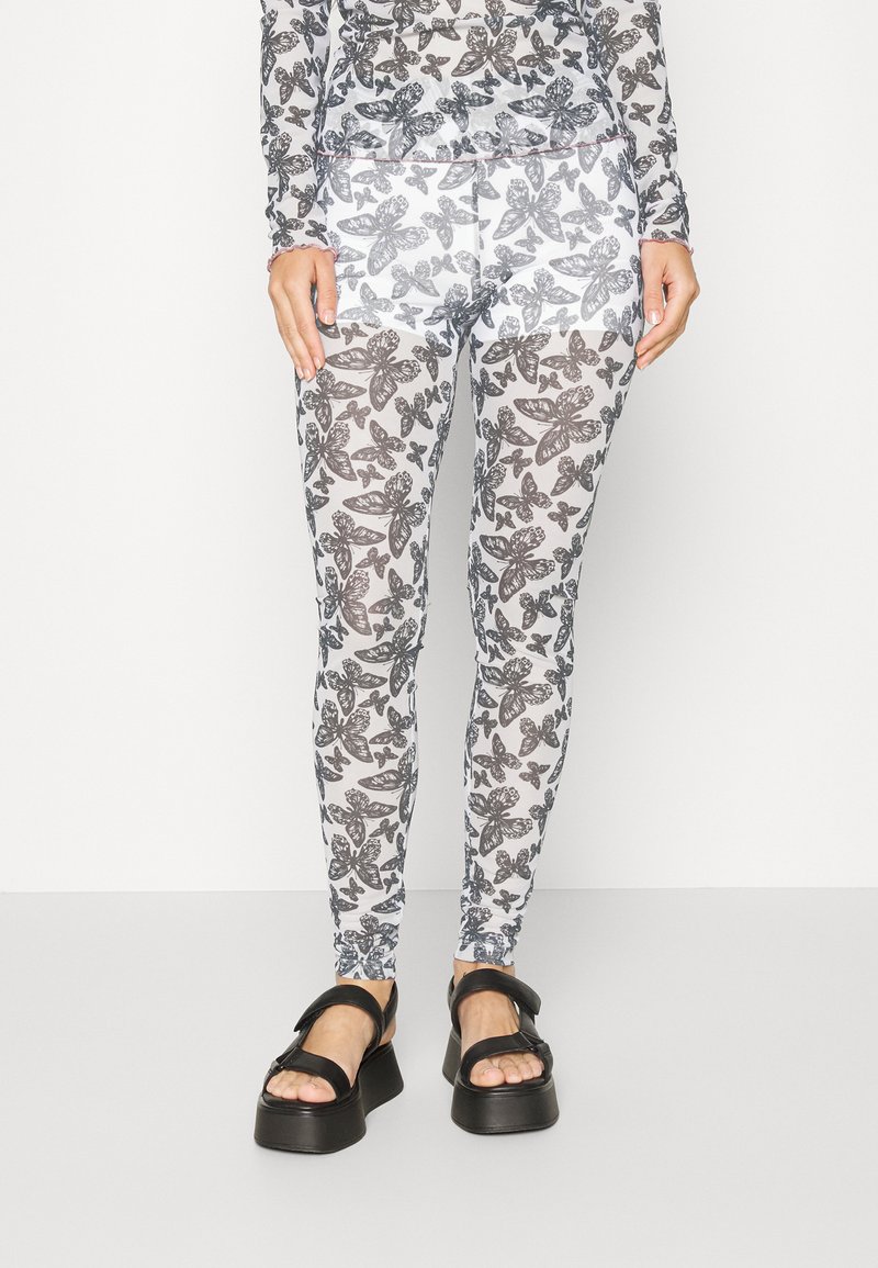 Leggings com padrão de borboletas em preto e branco feitos de tecido transparente, com um formato justo e faixa elástica na cintura. Combinados com sandálias pretas robustas.