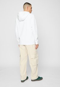 Felpa bianca con cappuccio regolabile, abbinata a pantaloni cargo beige con tasche laterali e sneakers verdi con suole texturate.