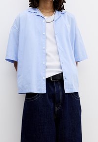 Camicetta a righe blu chiaro a maniche corte con chiusura a bottoni e texture morbida, abbinata a pantaloni larghi in denim scuro e cintura nera.