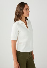 Polo bianco a maniche corte con un motivo a coste testurizzato e colletto a V. Abbinato a pantaloni verde oliva, visto di lato.