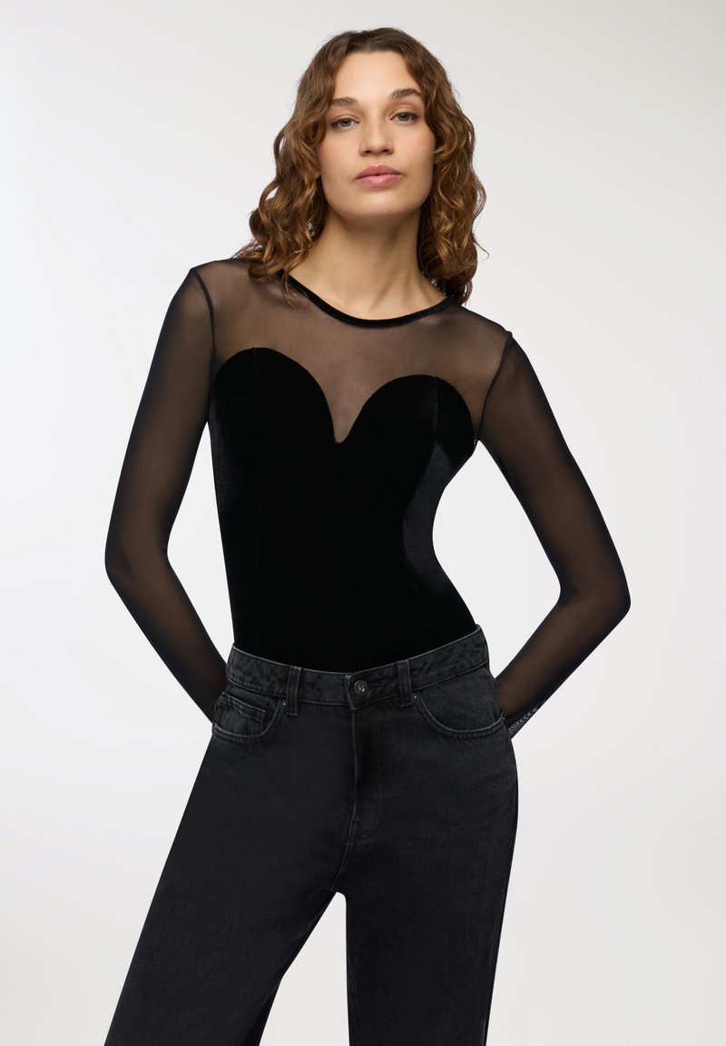 Body nero con scollatura in rete trasparente e maniche lunghe; presenta un'apertura a forma di cuore nella parte frontale; abbinato a jeans scuri.