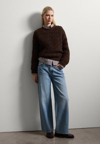 Brauner strukturierter Strickpullover über einem karierten Hemd, kombiniert mit hellblauen weiten Jeans und dunklen Slippern vor einem neutralen Hintergrund.