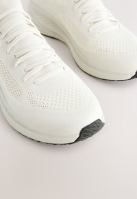 Chaussures de sport blanches avec tige en mesh et tissu, bout rond, lacets plats, semelle texturée, et un accent en caoutchouc noir sur le bout.