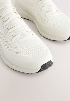 Weiße Sportschuhe mit Mesh- und Stoffschaft, rundem Zehenbereich, flachen Schnürsenkeln, strukturierter Sohle und einem schwarzen Gummieinsatz an der Spitze.
