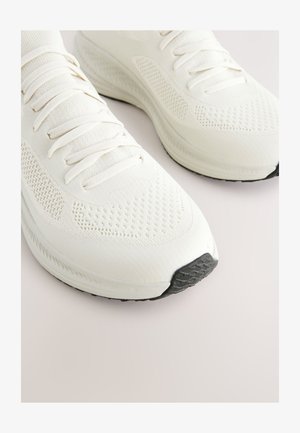 Weiße Sportschuhe mit Mesh- und Stoffschaft, rundem Zehenbereich, flachen Schnürsenkeln, strukturierter Sohle und einem schwarzen Gummieinsatz an der Spitze.