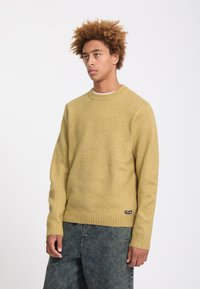 Jeune homme aux cheveux bouclés portant un pull en maille jaune moutarde et un jean gris foncé délavé, debout devant un fond clair uni.