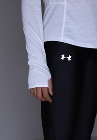 Biała bluzka z długim rękawem z delikatnymi detalami szycia, w zestawieniu z czarnymi legginsami z odblaskowym logo Under Armour na udzie.