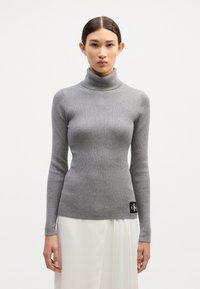 Grau gerippter Rollkragenpullover mit langen Ärmeln, taillierter Passform und einem schwarzen Markenpatch auf der unteren rechten Seite. Glatte Textur, dehnbare Stoffqualität.