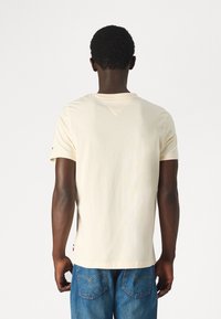 Hombre con el cabello rizado corto de pie de espaldas, vistiendo una camiseta plana color beige claro y jeans azules contra un fondo blanco.