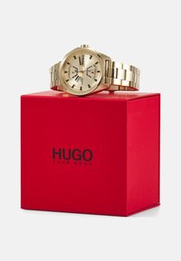 Reloj de oro con una pulsera metálica con eslabones, que presenta una esfera redonda y tres subdiales. Presentado en una caja cuadrada roja con un logo negro.