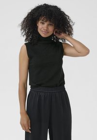Kvinde med krøllet hår iført sort ærmeløs turtleneck top og sorte vide bukser, stående foran en ensfarvet baggrund.