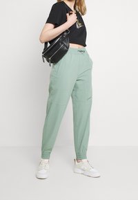 Kvinna i mintgröna Nike-joggers, vita sneakers, svart kortärmad Nike-t-shirt och svart axelväska stående mot en neutral bakgrund.