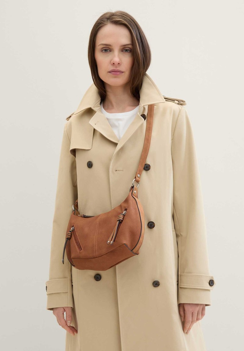 Femme portant un trench beige et une chemise blanche, portant un sac à bandoulière en cuir marron avec des détails à fermeture éclair
