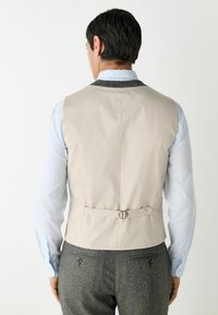 Beige vest with a smooth texture, rear buckle detail, and gray fabric accents, worn over a light blue shirt and gray patterned trousers.