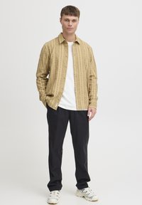 Camisa de botones de rayas marrón, camiseta blanca debajo, pantalones negros y zapatillas blancas. La camisa presenta patrones verticales y un ajuste relajado.