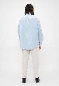 Hombre con rastas que lleva una camisa de manga larga de color azul claro con un patrón, pantalones beiges y zapatos negros, de pie de espaldas frente a un fondo blanco.
