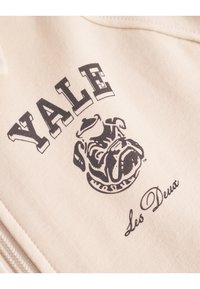 Cremefarget genser med svart trykt "Yale"-logo og bulldog-grafikk, med "Les Deux" under i kursiv skrift. Mykt bomullsmateriale.