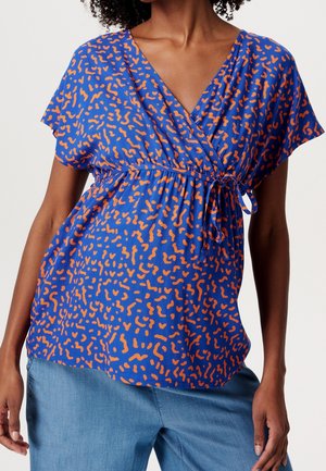 Mujer con blusa cruzada azul con estampado abstracto en naranja, atada a la cintura, combinada con vaqueros de mezclilla azul claro.